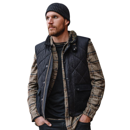 A man wearing a black quilted sleeveless vest with a high collar and front pockets, paired with a camouflage print shirt and a beanie.