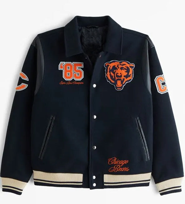 A Chicago Bears varsity jacket in navy blue with black leather-look sleeves, featuring orange and white embroidered details including the team bear logo, '85' on the chest, and 'Chicago Bears' script on the lower front.