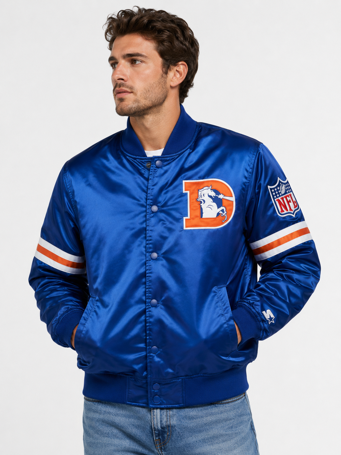 A man wearing a blue satin jacket with Denver Broncos graphics, including a large 'D' logo on the chest and an NFL shield on the sleeve, photographed against a light gray studio background.