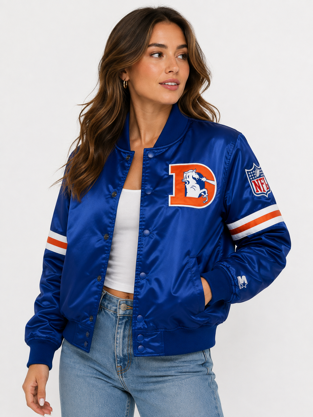 A woman wearing a royal blue satin bomber jacket with a large orange and white letter 'D' on the chest, representing the Denver Broncos, paired with a white crop top and light wash jeans against a plain white studio background.