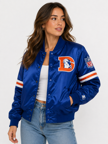 A woman wearing a royal blue satin bomber jacket with a large orange and white letter 'D' on the chest, representing the Denver Broncos, paired with a white crop top and light wash jeans against a plain white studio background.