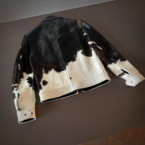 A woman wearing a cow-print jacket, cowboy hat, white crop top, and dark jeans stands in a bedroom setting, holding her cowboy hat.
