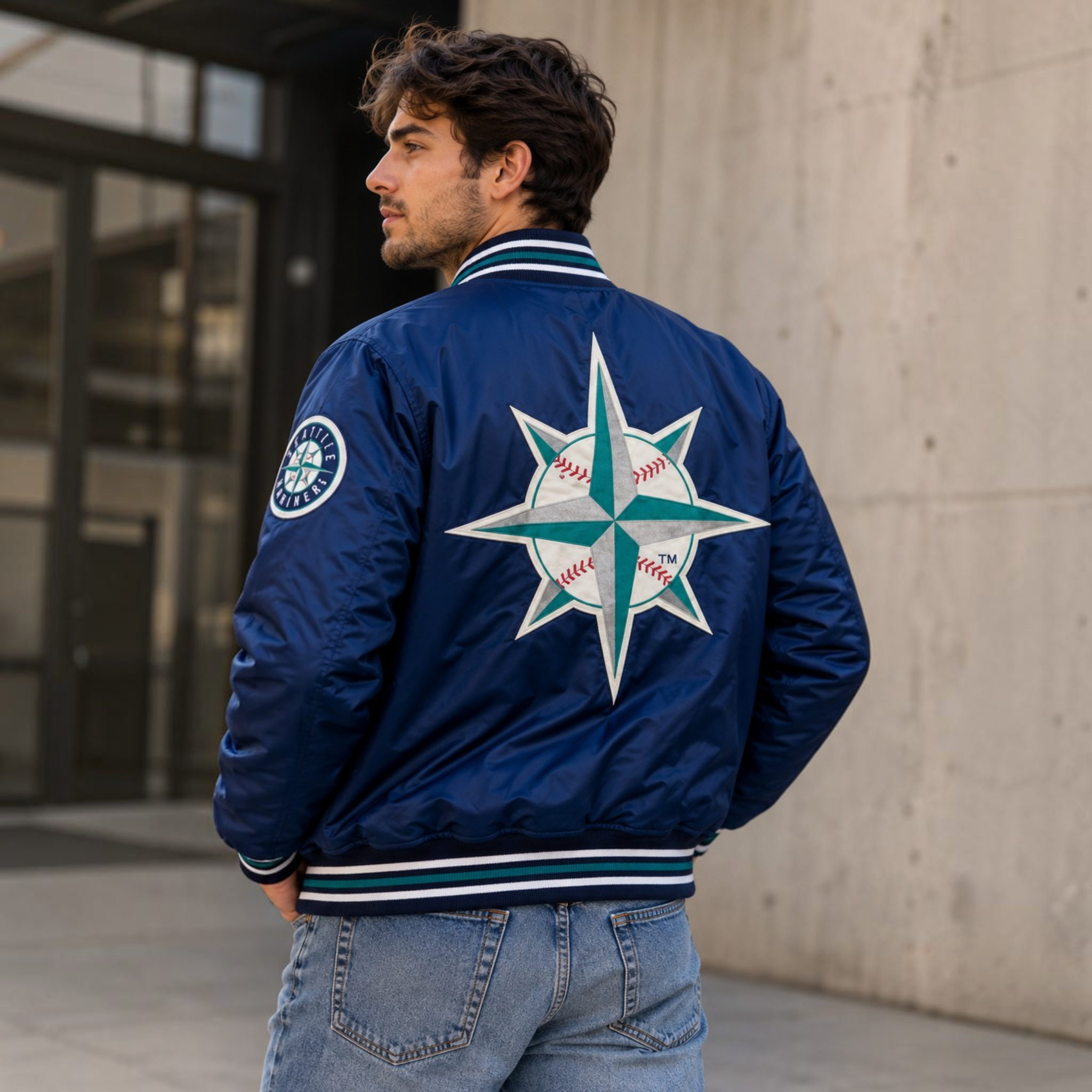 A man wearing a navy blue satin bomber jacket with a large Seattle Mariners baseball compass logo on the back, photographed outdoors against a concrete wall.