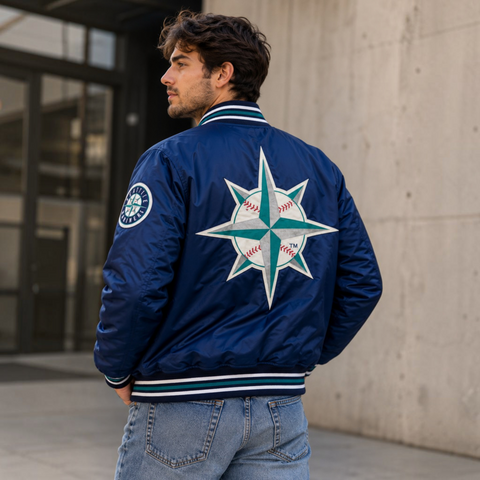 A man wearing a navy blue satin bomber jacket with a large Seattle Mariners baseball compass logo on the back, photographed outdoors against a concrete wall.