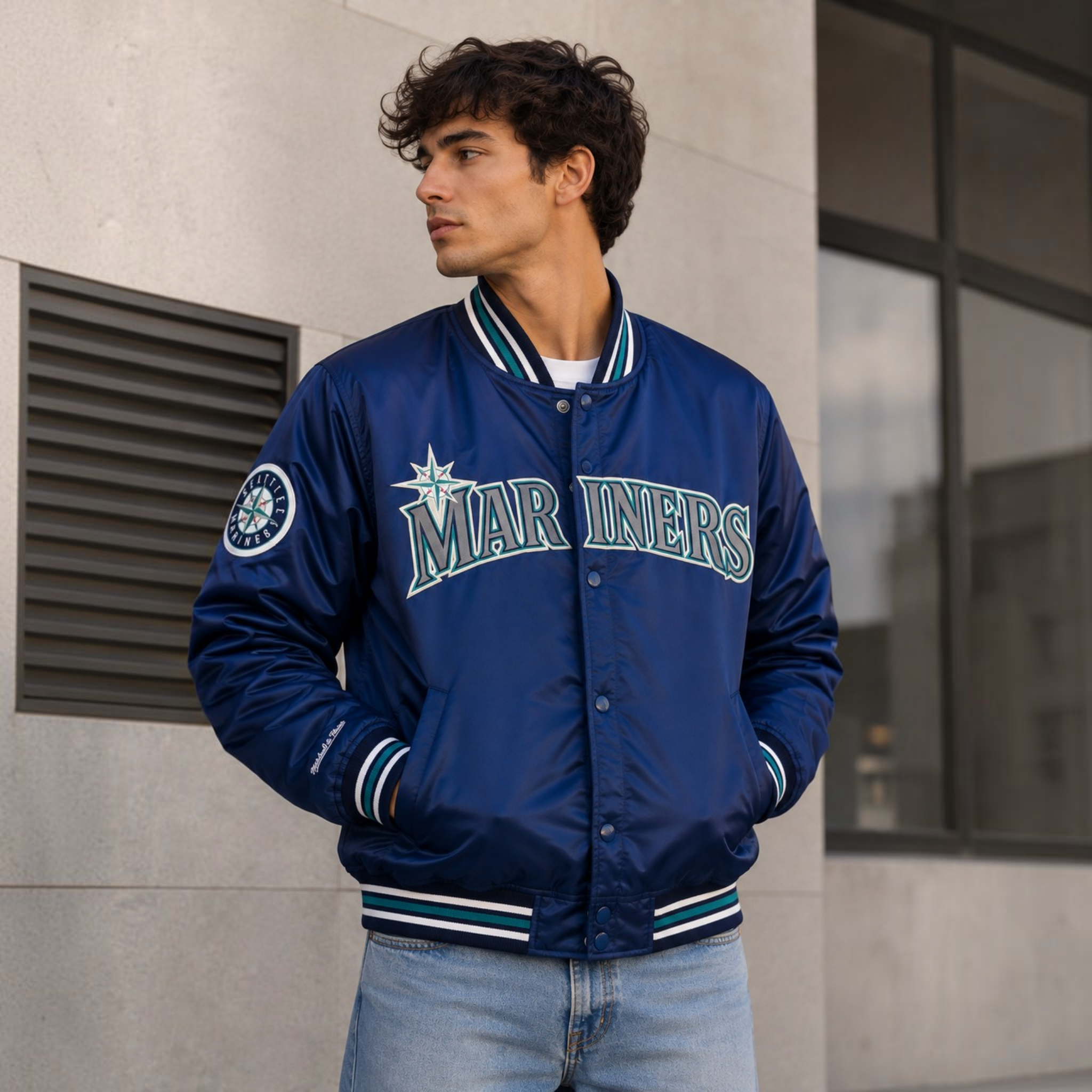 A man wearing a navy blue Seattle Mariners varsity jacket with 'MARINERS' written across the chest, paired with light blue jeans, standing outdoors against a concrete wall.