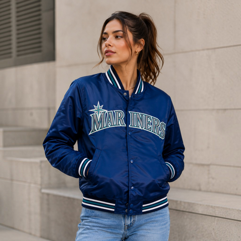 A woman wearing a navy blue Seattle Mariners varsity jacket with white and green striped ribbed trim, standing against a light gray stone wall.