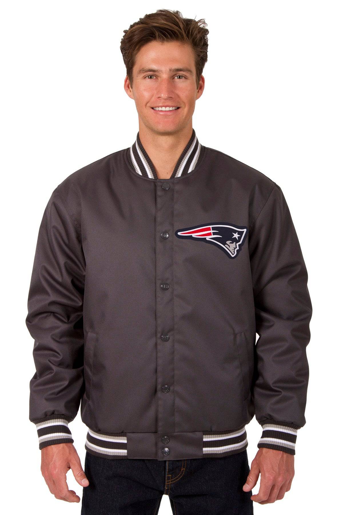 A man wearing a dark gray satin varsity jacket with the New England Patriots logo on the chest, featuring striped ribbed collar, cuffs, and hem, set against a plain white background.