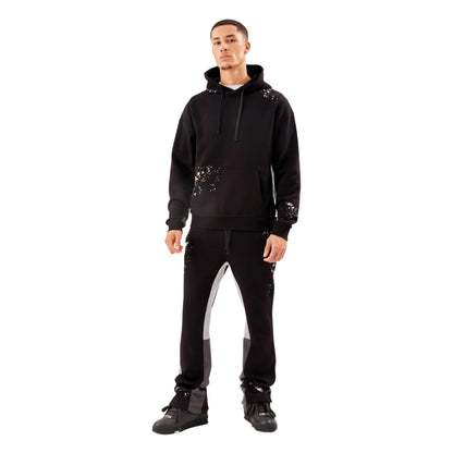 A male model wearing a black tracksuit with a unique paint smear pattern, featuring a hooded jacket and matching pants.