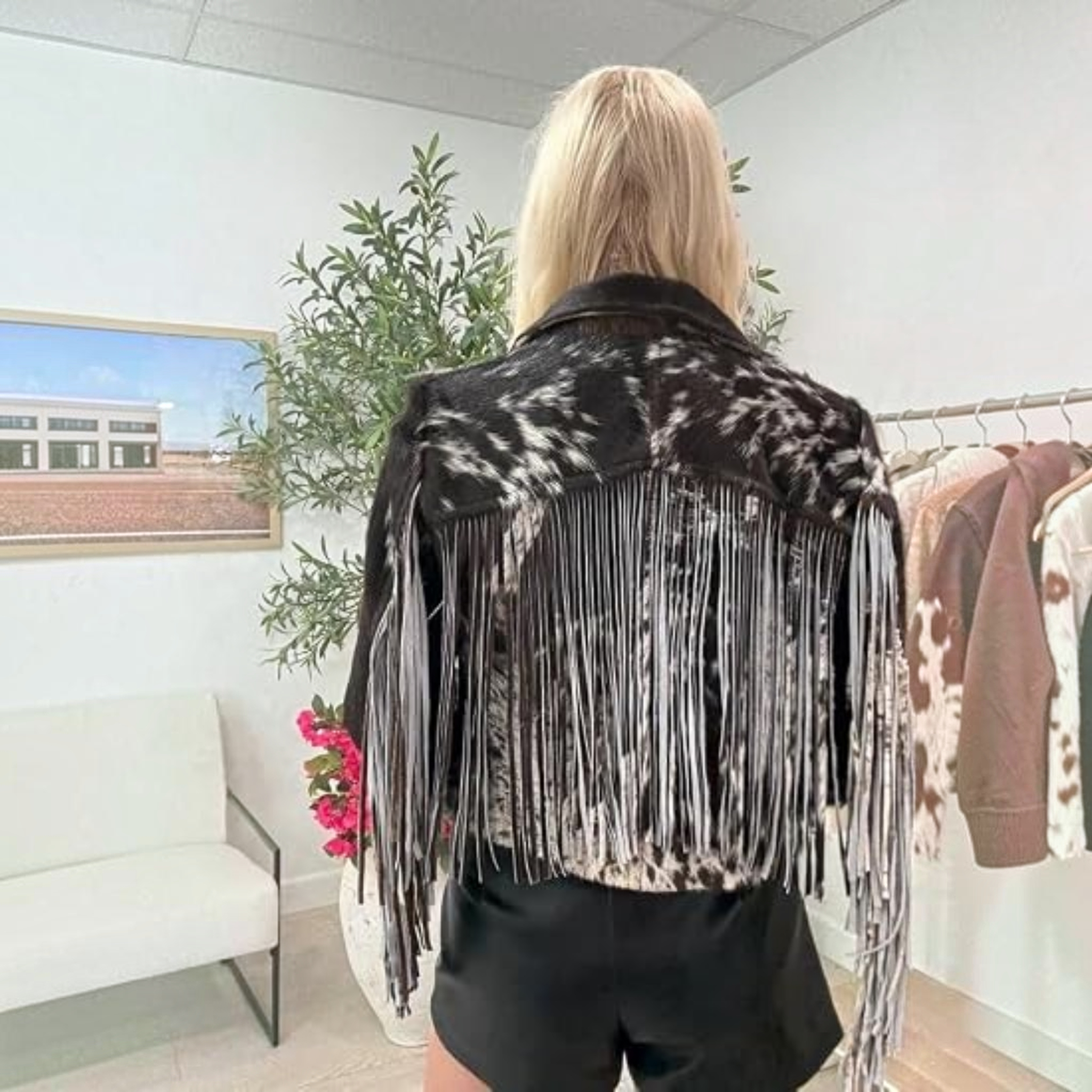 A woman wearing a black and white cow-print cropped jacket, a brown cowboy hat, a white top, and dark jeans, standing in a bedroom setting.