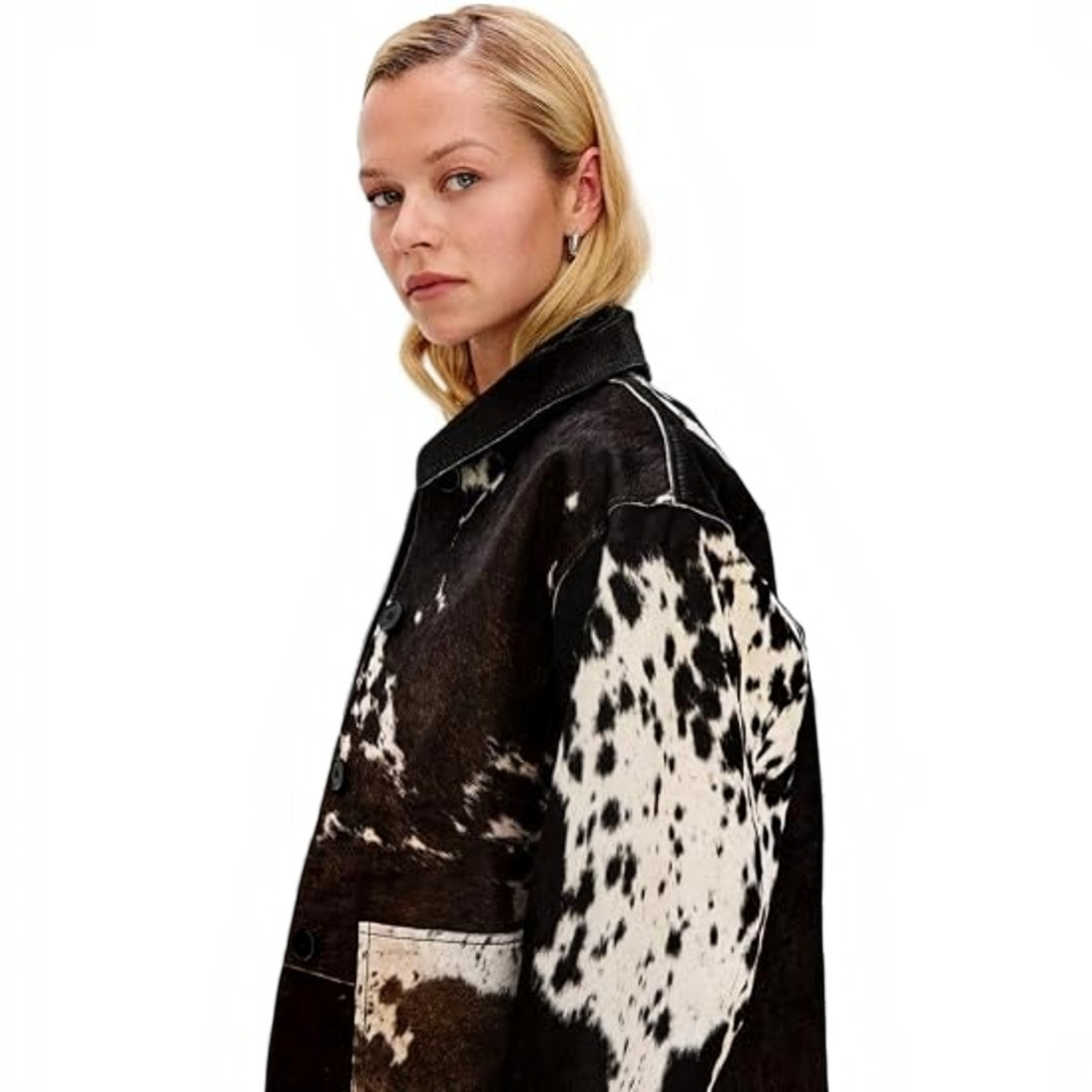 A woman wearing a black and white cow-print cropped jacket, a brown cowboy hat, a white top, and dark jeans, standing in a bedroom setting.