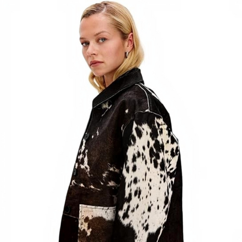 A woman wearing a black and white cow-print cropped jacket, a brown cowboy hat, a white top, and dark jeans, standing in a bedroom setting.