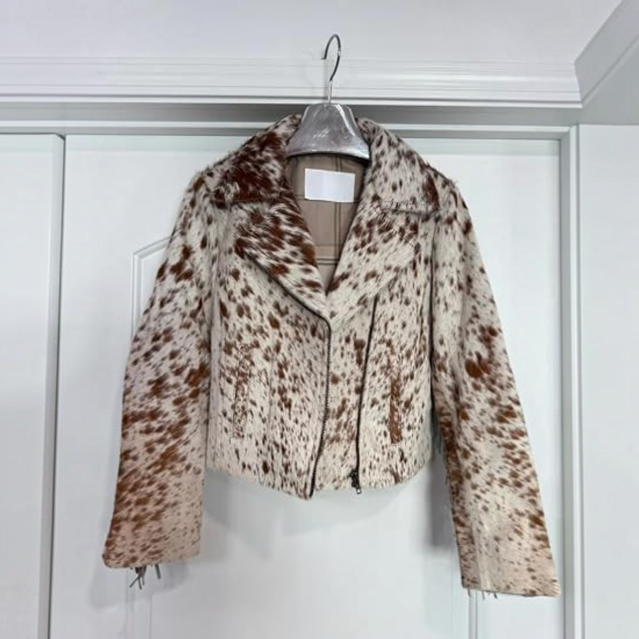 A cropped faux fur motorcycle jacket with a brown and cream cow print pattern, hanging on a silver hanger against a white paneled door.