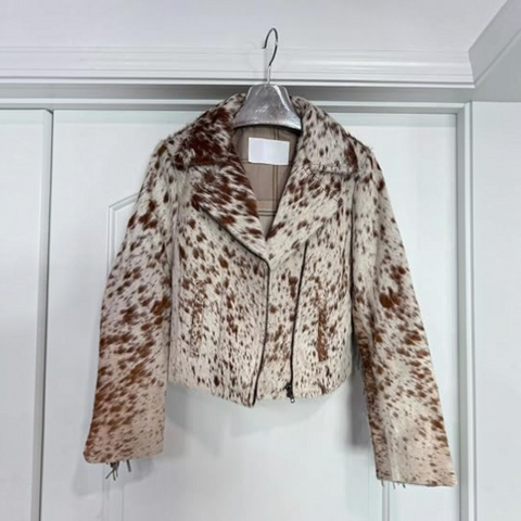 A cropped faux fur motorcycle jacket with a brown and cream cow print pattern, hanging on a silver hanger against a white paneled door.