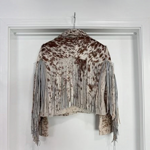 A person stands on outdoor steps in front of a brown door and curtain, wearing a brown and white cow-print jacket over a cream top, olive wide-leg jeans, and holding a pink textured handbag.