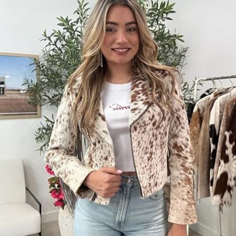 A woman modeling a cropped, long-sleeve faux fur jacket with a brown and beige animal print pattern, worn over a white graphic t-shirt and light blue jeans in a boutique setting.