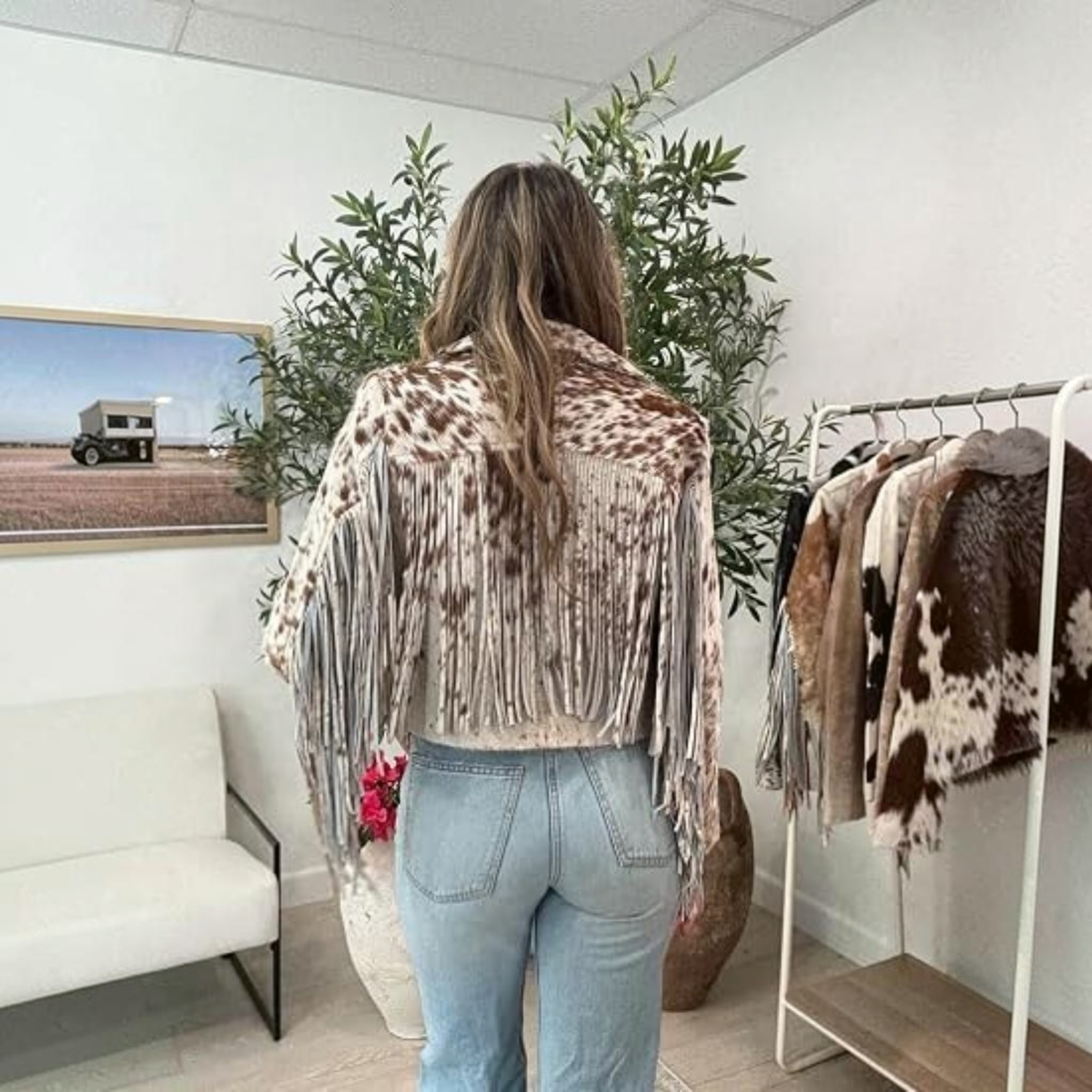 A person wearing a brown and white cow-print faux fur jacket stands in front of a fruit stand at a street market, looking at red apples and yellow citrus fruits.