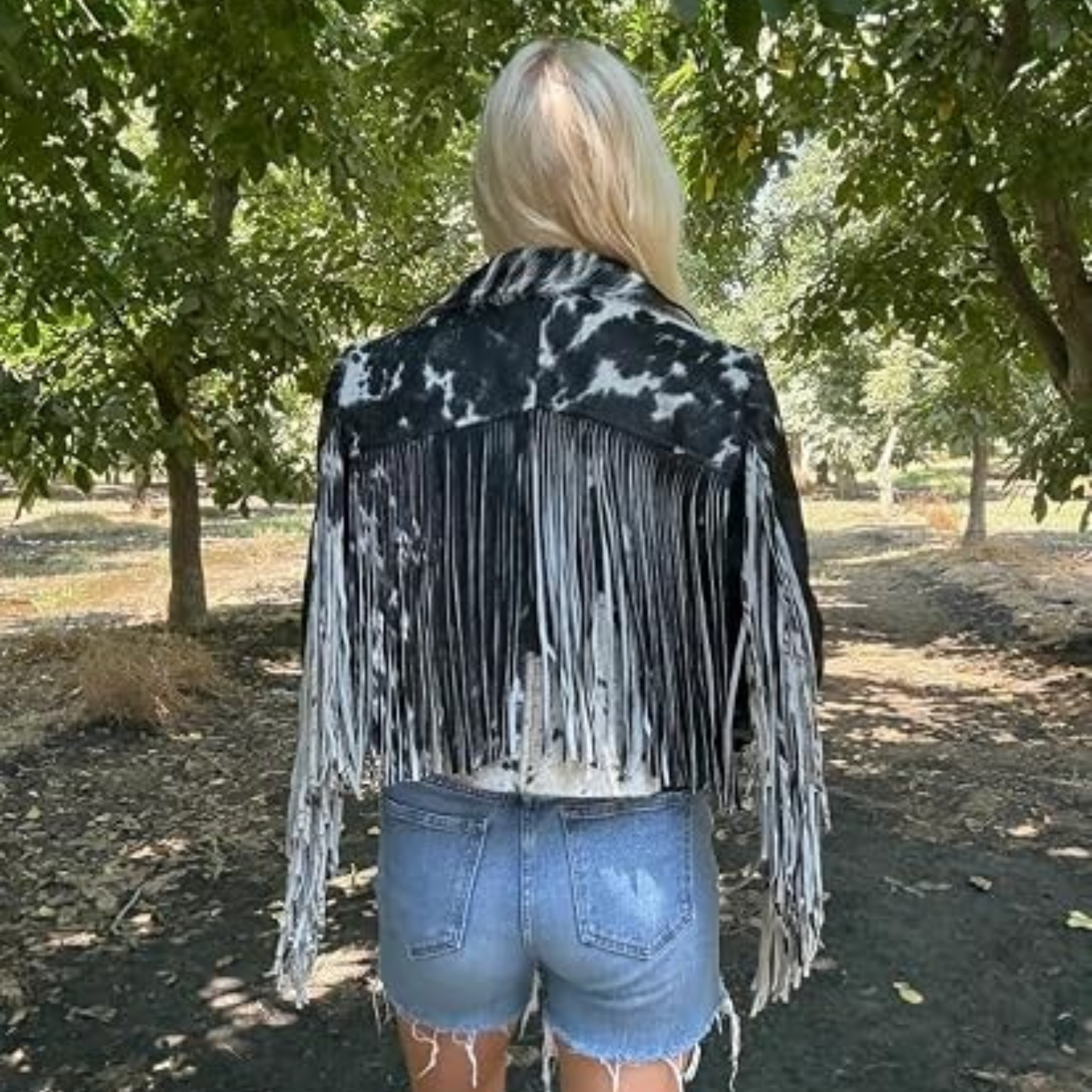 A woman wearing a black and white cow-print fringed jacket and a black cowboy hat, shown against a plain white background.