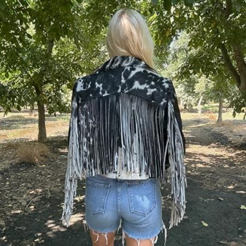 A woman wearing a black and white cow-print fringed jacket and a black cowboy hat, shown against a plain white background.