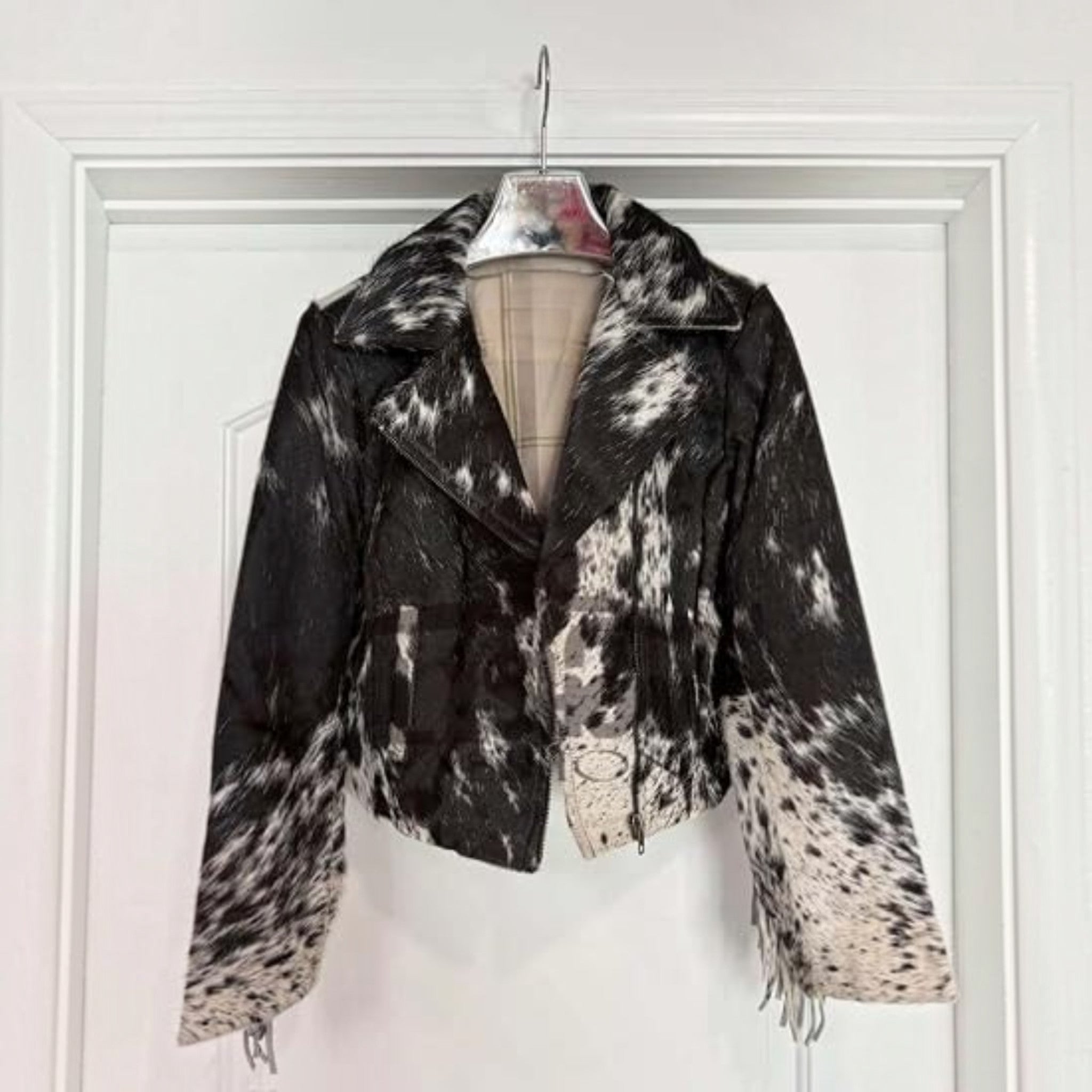 A cropped motorcycle jacket with a black and white cow print pattern, hanging on a hanger against a white paneled door.