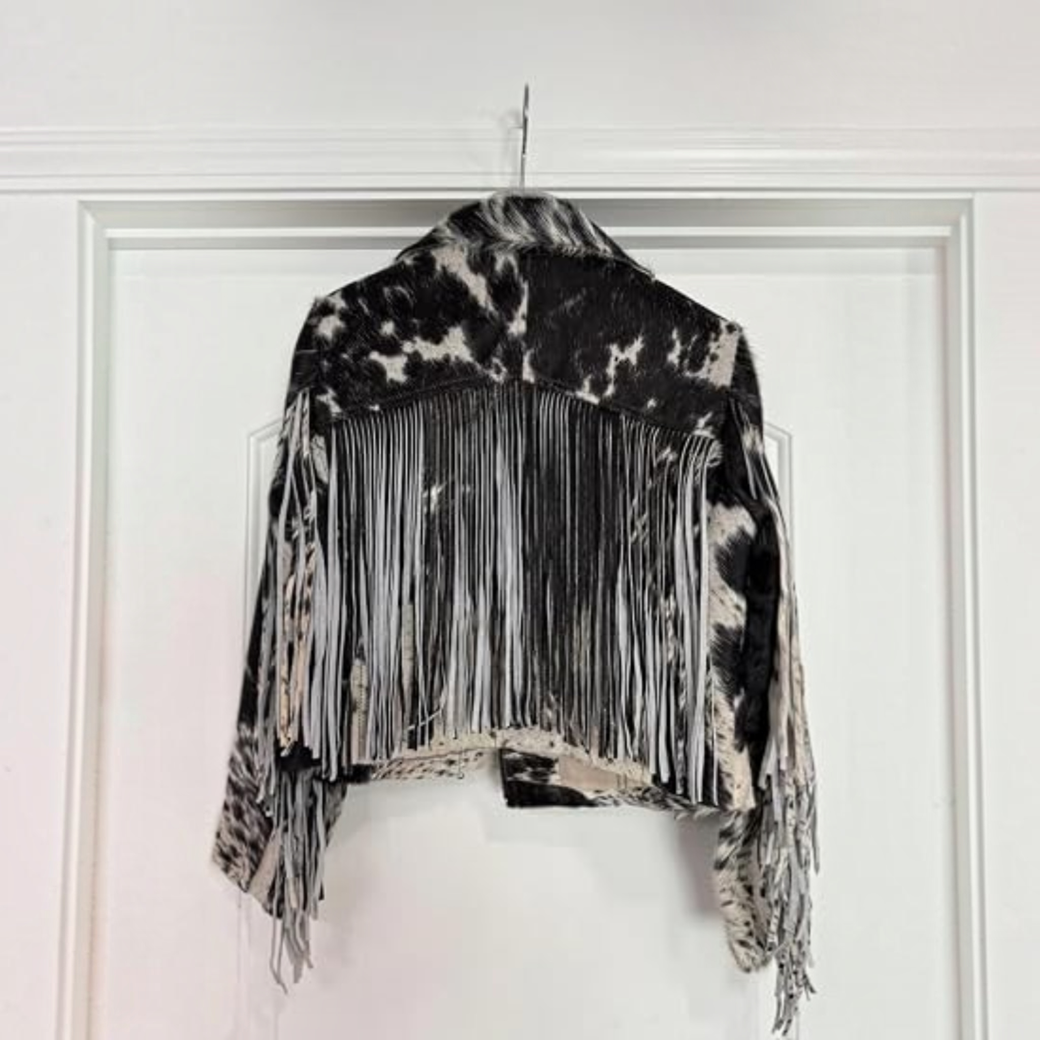 A woman modeling a black and white cow-print fringed jacket with a black cowboy hat and black faux-leather pants, set against a plain white background.