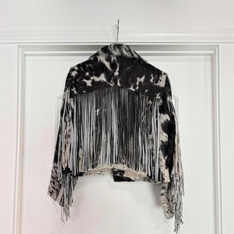 A woman modeling a black and white cow-print fringed jacket with a black cowboy hat and black faux-leather pants, set against a plain white background.