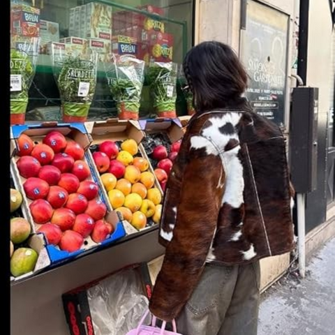 A person wearing a brown and white cow-print faux fur jacket stands at an outdoor fruit stand next to a shop window displaying packaged greens.