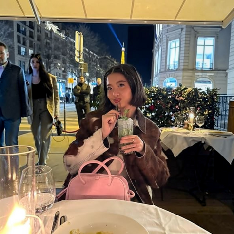 A woman sits at an outdoor restaurant table at night, holding a drink with a straw. She is wearing a brown and white patterned coat and a pink handbag sits in front of her.