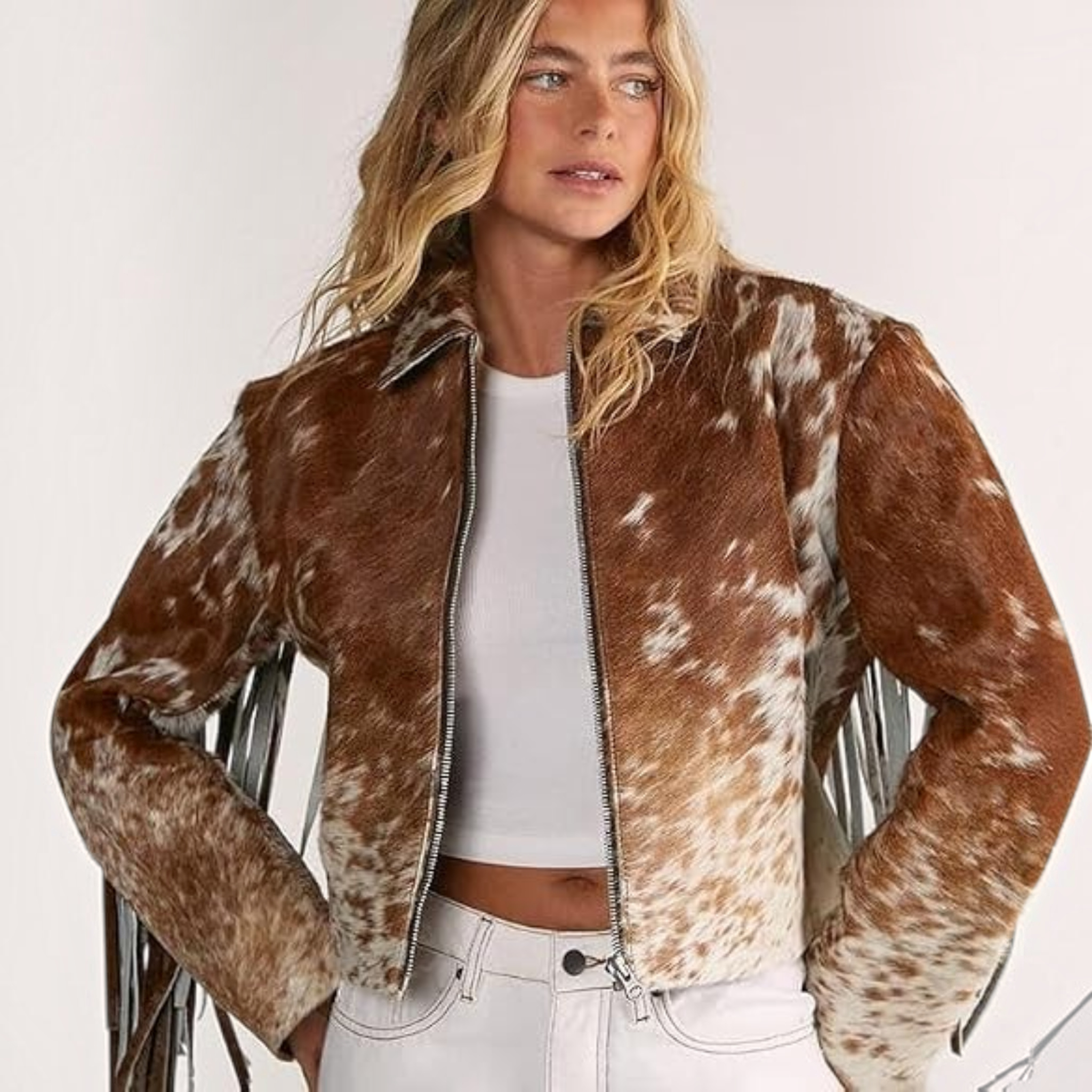 A woman wearing a brown and white cow-print faux fur jacket with fringe details, styled over a white crop top and white jeans against a plain light background.