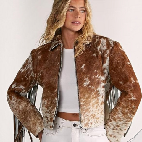 A woman wearing a brown and white cow-print faux fur jacket with fringe details, styled over a white crop top and white jeans against a plain light background.
