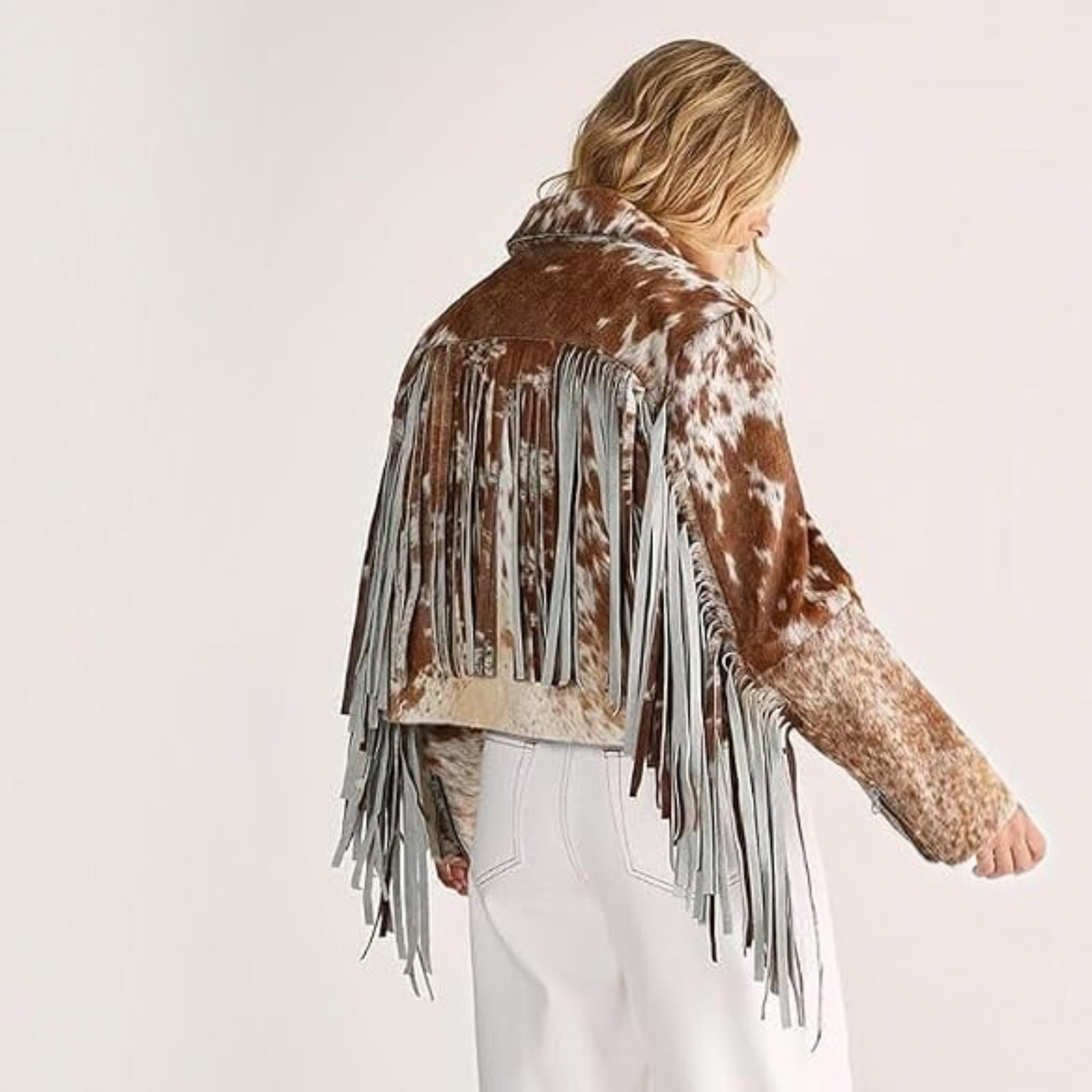 A woman shown from the back wearing a brown and white cow-print fringed jacket with white pants, photographed against a plain light background.