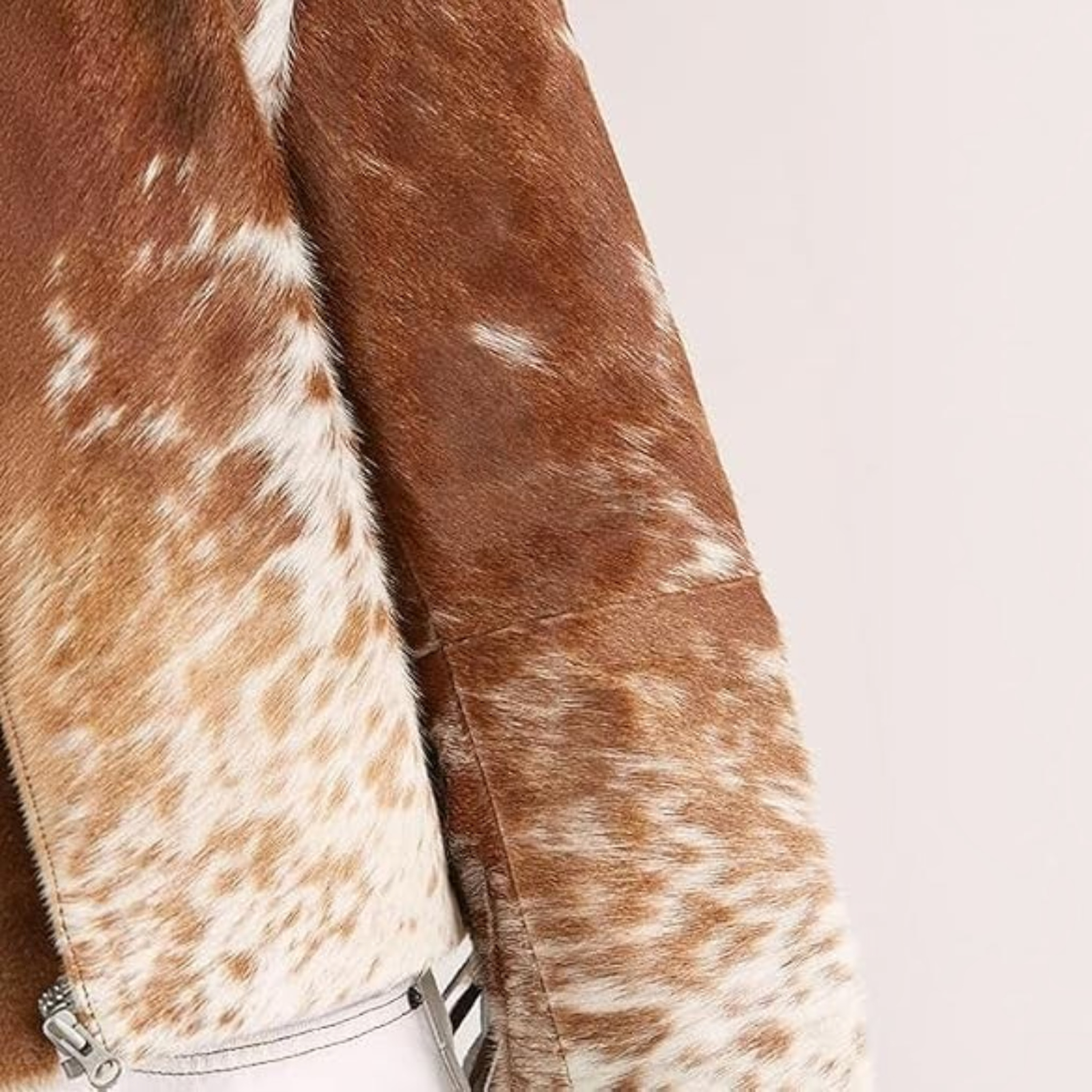 Close-up of a brown and white cowhide jacket or coat with a visible silver zipper and a small portion of a white garment underneath, against a plain light background.