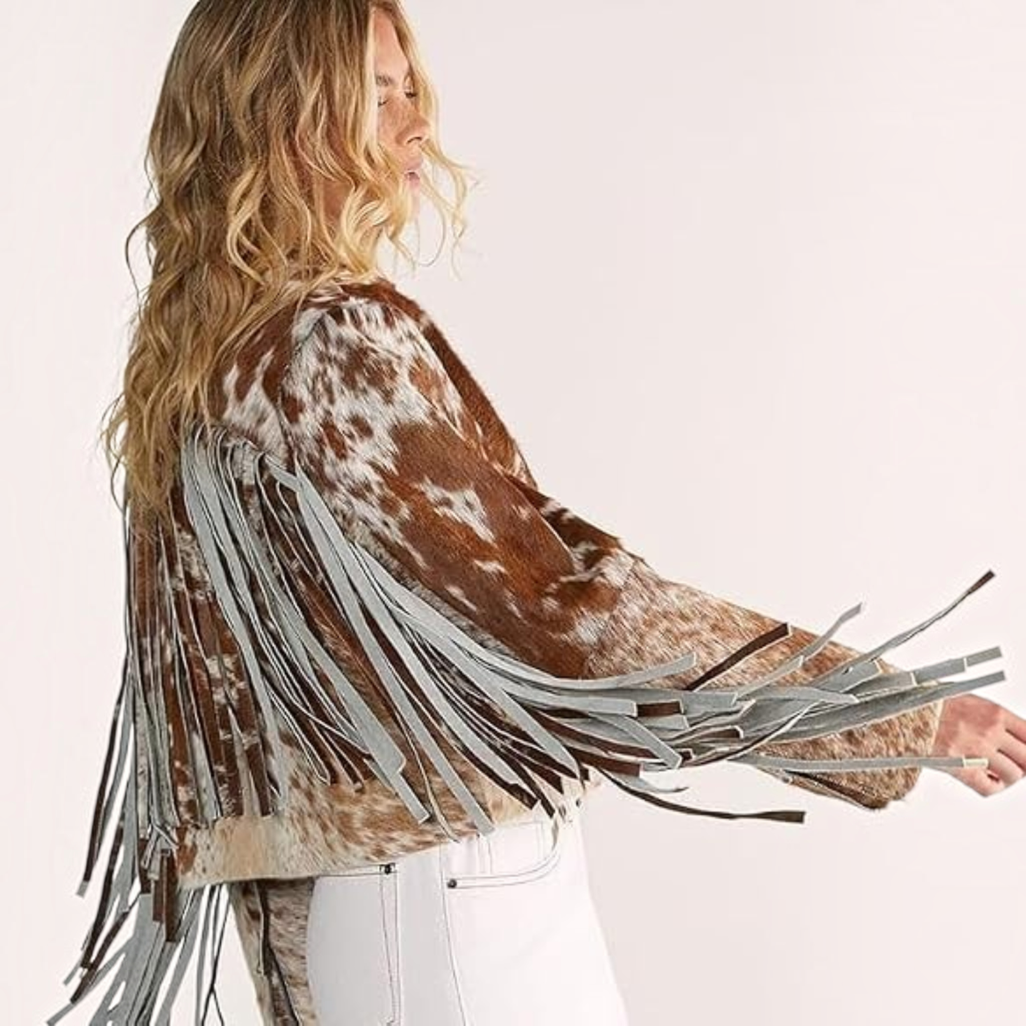 A woman shown from the side and back wearing a brown and white cow-print fringed jacket with long gray fringe, paired with white pants, against a plain light background.