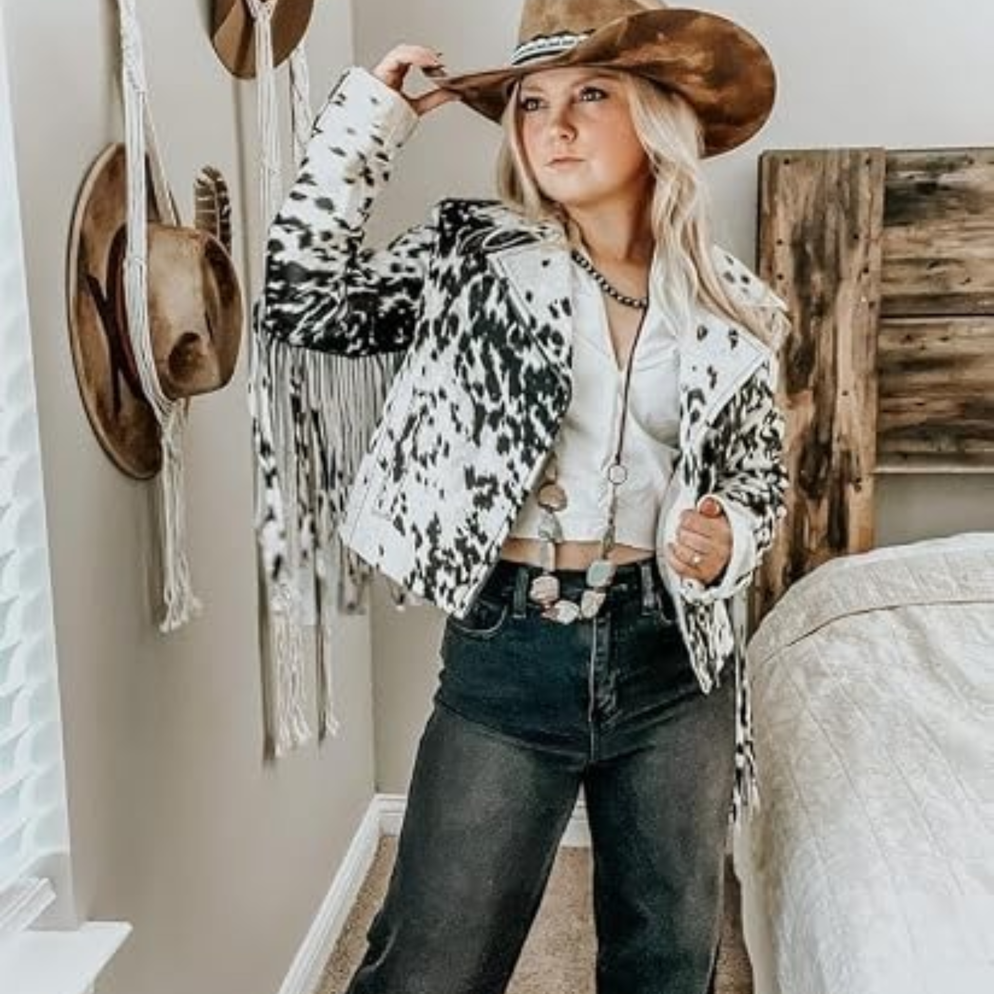 A woman wearing a cow-print fringed jacket, a brown cowboy hat, a white crop top, and dark jeans, posing in a bedroom setting with a rustic wooden headboard and wall decorations.