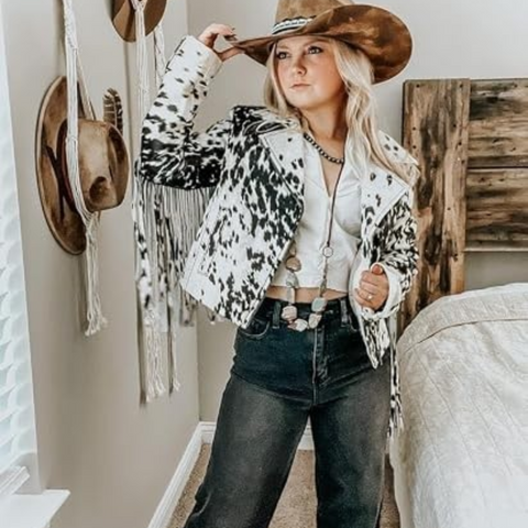 A woman wearing a cow-print fringed jacket, a brown cowboy hat, a white crop top, and dark jeans, posing in a bedroom setting with a rustic wooden headboard and wall decorations.