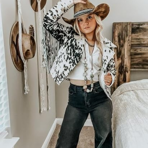 A woman modeling a black and white cow-print fringed jacket with a black cowboy hat and black faux-leather pants, set against a plain white background.