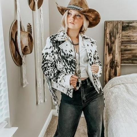 A woman wearing a black and white cow-print cropped jacket, a brown cowboy hat, a white top, and dark jeans, standing in a bedroom setting.