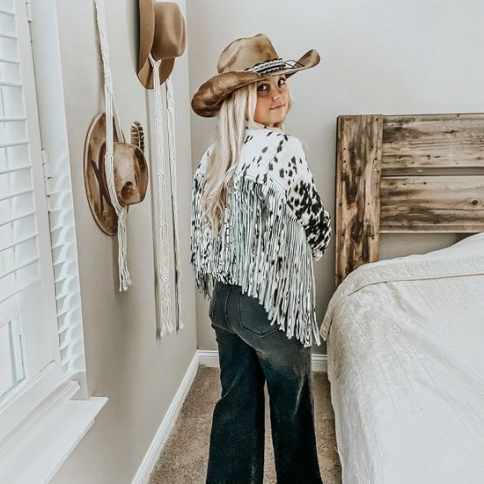 A woman stands in a bedroom wearing a brown cowboy hat, a white fringed poncho with black spots, and dark jeans, looking back over her shoulder. A bed with white bedding and a rustic wooden headboard is visible next to her, along with wall-mounted hats and macrame decor.