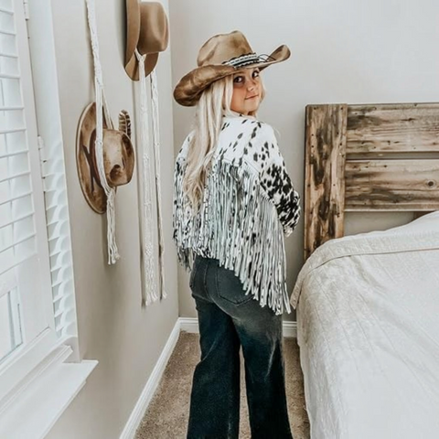 A woman stands in a bedroom wearing a brown cowboy hat, a white fringed poncho with black spots, and dark jeans, looking back over her shoulder. A bed with white bedding and a rustic wooden headboard is visible next to her, along with wall-mounted hats and macrame decor.