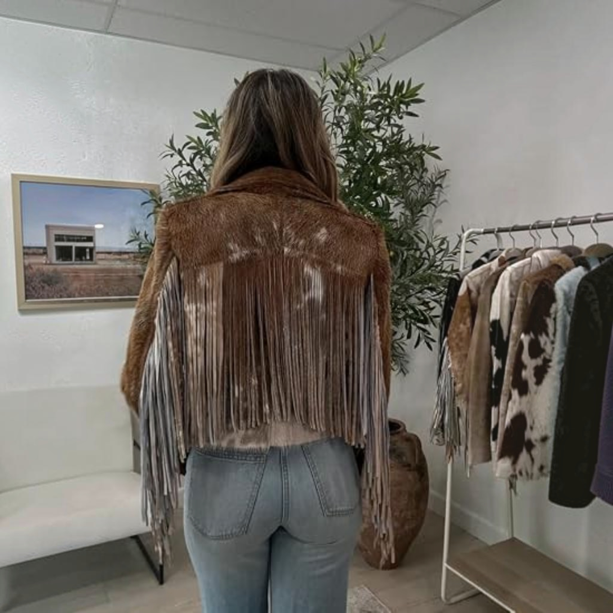 A woman wearing a cow-print jacket, cowboy hat, white crop top, and dark jeans stands in a bedroom setting, holding her cowboy hat.