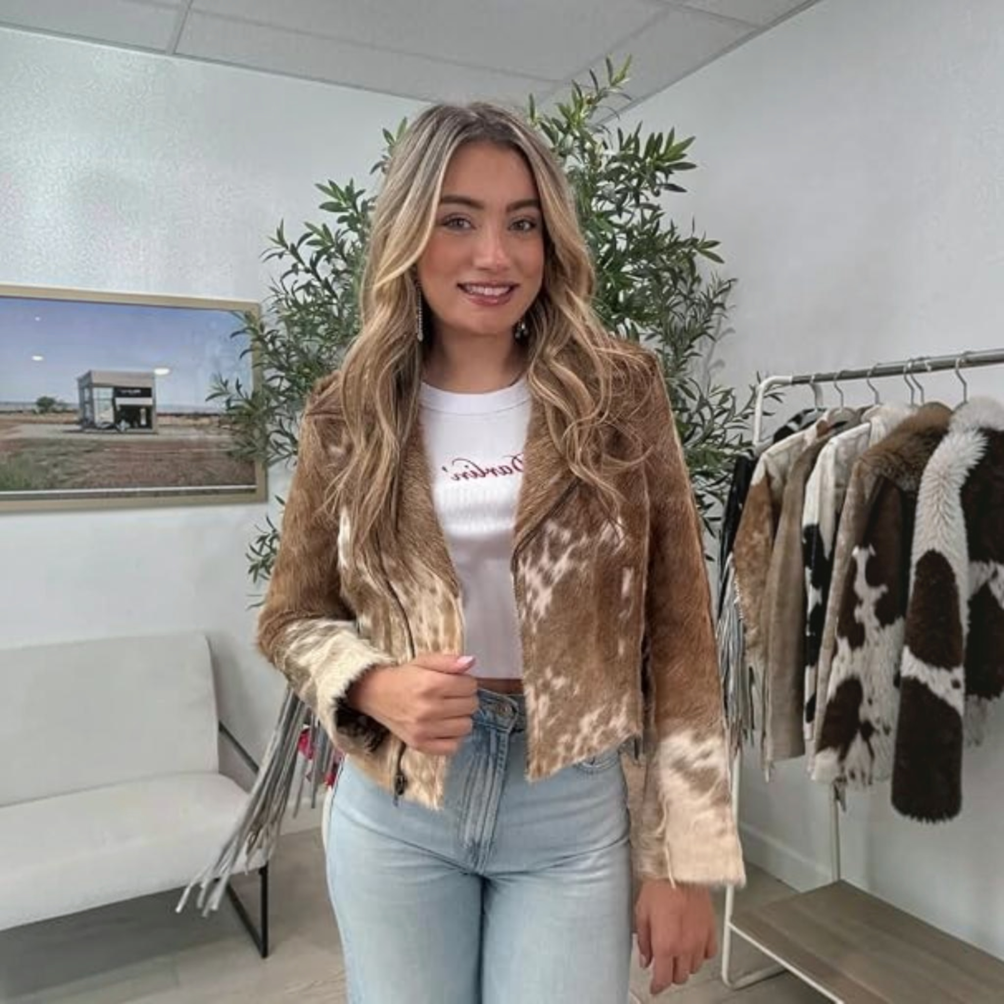 A woman wearing a black and white cow-print cropped jacket, a brown cowboy hat, a white top, and dark jeans, standing in a bedroom setting.