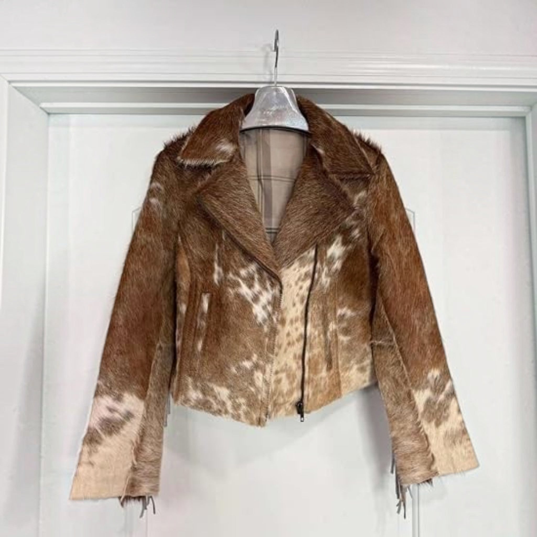 A cropped faux fur jacket with a brown and beige cow-print pattern, hanging on a clear plastic hanger against a white door.